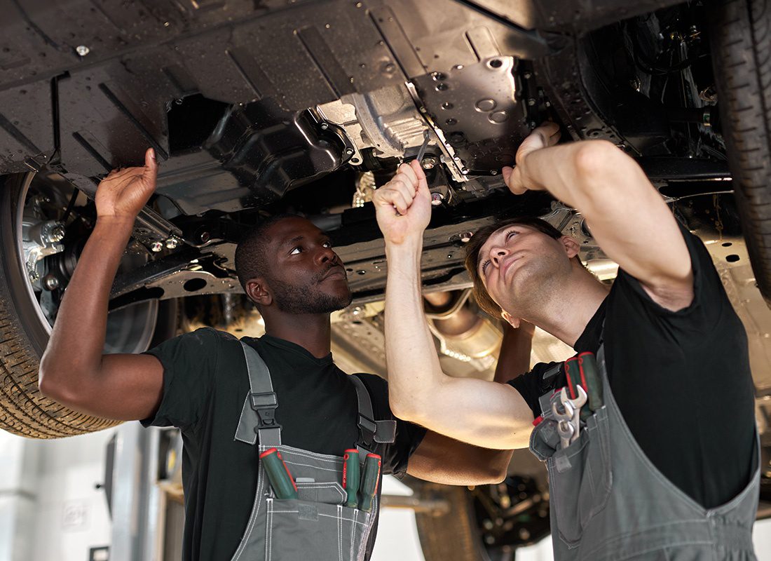 Business Insurance - Two Auto Technicians Wearing Black and Grey Uniforms Work on the Under Carriage of a Car